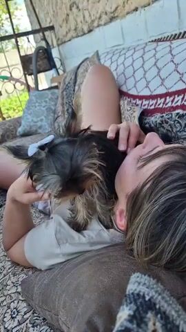 A person lies on a patterned couch outdoors with a small dog. The person's head is tilted back, resting on a cushion, and their arm is around the dog. The dog, a Yorkshire Terrier, has a small white bow in its fur and is nuzzling the person's face. The dog briefly sticks out its tongue. The background shows a textured wall and foliage. The overall impression is one of affection between the person and the dog.