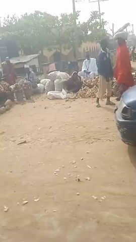 The video shows a scene of a bustling outdoor market in a rural area. The ground is sandy and dusty. In the first few shots, we see a dark-colored car parked, and then driving slowly. People are seen milling about, some wearing traditional clothing. There are large piles of what appears to be produce, like potatoes or yams, in buckets and sacks. A security guard is visible in the crowd. Later, a large blue MAN truck passes by. The overall atmosphere suggests a typical market day in a small town.
