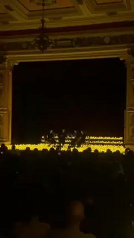 The scene captures a live performance viewed from the audience. A string quartet is playing on a stage illuminated by many small, yellow lights, resembling candles. The stage is framed by a grand, ornately decorated proscenium arch with a dark backdrop. The audience is silhouetted in the foreground, with many individuals holding up phones, presumably to record or photograph the performance. The overall atmosphere appears elegant and intimate.