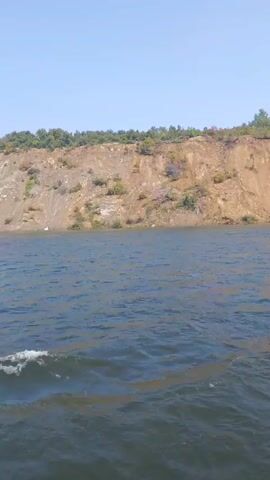 The video shows a body of water with a steep, eroded cliff face on the far side. The cliff is covered in sparse vegetation and appears to be made of sandy soil. The water is choppy, indicating some wind or current. On the right side of the frame, part of a structure is visible, possibly the side of a boat or dock, with a white railing. The sky is clear and blue. The camera appears to be moving slowly, likely on the water.