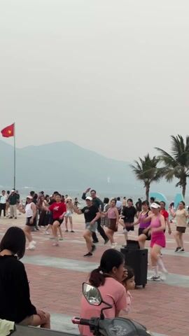 The video shows a group of people participating in a dance workout in what appears to be a public plaza near a beach. The dancers, of varying ages and dressed in casual exercise clothing, are following a routine led by an instructor. In the foreground, two women, one with a young child, are observing the group from a bench next to a moped. The plaza is paved with red and gray bricks, and palm trees dot the area. In the background, the ocean is visible, with mountains in the distance. The sky is overcast. A large "DA NANG" sign stands near the beach. People walking by are also visible in the background. A portable speaker is positioned near the center of the dance group, likely providing the music for the workout.