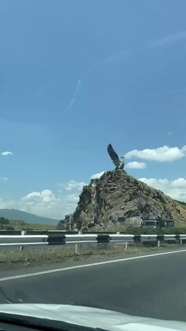The video shows a large metal sculpture of a bird with outstretched wings perched atop a rocky hill. The sculpture is silver and appears to be an eagle or hawk. The hill is covered with rocks and sparse vegetation. In the background, there is a blue sky with some white clouds and a contrail from an airplane. A highway with guardrails runs in front of the hill. There is a white truck and other vehicles visible on the road and near the base of the hill. The video appears to be filmed from a moving vehicle, as evidenced by the slight blur and perspective shifts. The overall impression is one of a roadside monument in a natural landscape.