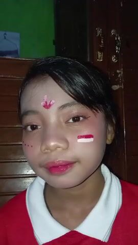 This is a close-up, medium-angle video of a young girl with dark hair and light skin. She is wearing a red shirt with a white collar and has elaborate face paint. The face paint includes a pink design on her forehead, red dots with glitter around her eyes and on her cheeks, and the Indonesian flag painted on her right cheek. Her lips are painted a glossy pink. The background appears to be indoors with wooden furniture and a dark doorway, suggesting a domestic setting. The lighting is somewhat dim, but her face is well-lit. The girl blinks and shifts her gaze slightly throughout the short clip, showing a neutral or slightly playful expression. The overall impression is that of a child participating in a cultural or celebratory event, possibly related to Indonesian Independence Day, given the flag imagery.