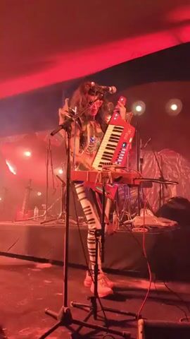 A performer wearing a futuristic, outlandish costume is singing and playing a keytar on a stage. The performer has long dark hair and is wearing elaborate shoulder pieces, a bejeweled top, and striped pants. The stage is dimly lit with red and blue lights. A microphone stands in front of the performer, and a drum set is visible in the background. Smoke or haze hangs in the air, adding to the atmospheric effect.