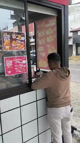 A person wearing a light brown hooded sweatshirt and light-colored pants is standing in front of a window at what appears to be a fast-food establishment. The person is holding several banknotes and appears to be in the process of paying or counting money. Through the window, a menu board displaying images of fried chicken and prices is visible. The environment outside the window suggests an urban setting with buildings and a street. The window itself has a tiled frame and a black trim.