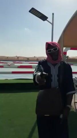 The video shows a man standing next to a water-based obstacle course. The man is wearing traditional Arab clothing including a red and white keffiyeh headdress, sunglasses, and a long dark blue tunic. He is also carrying a brown bag on his shoulder and holding a phone in his hand.

The obstacle course is set in a shallow body of water. It consists of red and white striped barriers creating a maze-like path. In the background, there's a desert landscape with sand dunes and some palm trees visible. The sky is clear and blue. A tall white light pole is also visible next to the man. The man briefly looks at the camera.