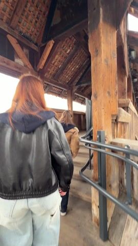 The camera moves along a wooden covered bridge with wooden beams and triangles of paintings above. People are walking along the bridge in both directions. Most of the people are wearing casual clothes like jackets and jeans. The camera is behind a red haired person in a black jacket and ripped jeans.