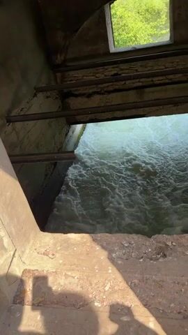 The video shows a close-up shot of turbulent, foamy water flowing forcefully through an industrial-looking opening. The opening appears to be part of a concrete structure with metal bars running across the top. Above the opening, a small, rectangular window reveals a bright green, leafy exterior, suggesting an outdoor environment. The water is a pale green color and churns vigorously as it passes through the channel. The perspective is from above and slightly to the side, looking down into the water flow. The overall impression is one of powerful water movement within a functional structure.