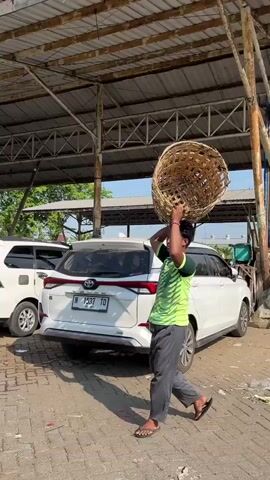 A young man walks under a covered structure, carrying a large woven basket on his head. He wears a green t-shirt and dark pants. He is walking to the right of the frame. To his left, a white SUV is parked. The ground is paved with brick-like stones. The environment appears to be an outdoor market or transportation hub. The lighting suggests it is daytime, with shadows cast by the structure and the man.