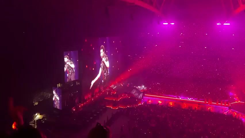 The video shows a concert performance taking place in a large, dimly lit arena. The stage is elaborate, featuring multiple screens displaying images of the performer, who is dressed in a dark, sparkly outfit. The performer is seen on stage and on the large screens, gesturing and singing. The audience is visible in the foreground and background, illuminated by the stage lights and small lights held by concert-goers. The lighting is dramatic, with beams of purple and red light cutting through the smoky atmosphere. There are also numerous small, bright lights, possibly confetti or LEDs, scattered throughout the arena. The overall impression is one of an energetic and visually stimulating live musical event.