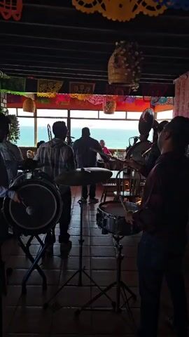 The video displays a group of musicians playing in a dimly lit room with a scenic ocean view. In the foreground, three musicians are visible. On the left, a musician is playing a large drum with a mallet. In the center, a musician stands behind a snare drum and cymbal, also holding drumsticks. To the right, another musician is playing a wind instrument. Behind the musicians, a man stands facing the ocean, which is visible through a large window. The room is decorated with colorful papel picado banners hanging from the ceiling. The overall atmosphere appears to be festive, possibly during a meal or celebration, given the presence of tables and chairs visible in the background.