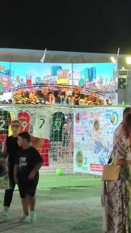 The video shows a night scene with a large digital screen displaying a cityscape and a spinning circular platform with people on it. In the foreground, there is a soccer goal with several jerseys hanging on it, and a green soccer ball is on the ground in front of the net. Several people are standing around the goal, including two children in the lower left and a woman in a patterned dress on the right. The atmosphere appears to be that of an outdoor event or festival.