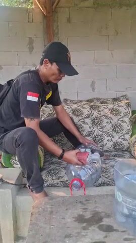 A person wearing a black baseball cap and a black t-shirt with an Indonesian flag patch is using an angle grinder to cut a large plastic water bottle. The person is sitting on a patterned cushion in an outdoor, semi-enclosed space with a concrete block wall in the background. There are other plastic water bottles visible.