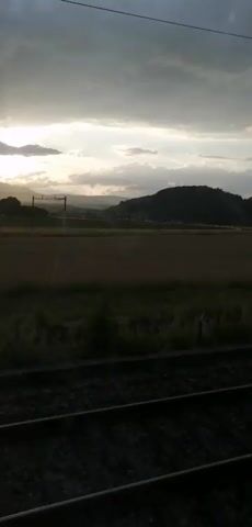 This media file captures a scenic view from a train window as it travels through Daillens, Switzerland. The scene depicts a rural landscape under a dramatic, cloudy sky, likely at dusk or dawn, given the soft, diffused sunlight breaking through the clouds.

The primary subjects are the natural elements of the landscape: rolling hills covered in dark vegetation in the distance, and expansive fields of farmland in the midground. The fields appear to be a mix of cultivated crops and grasslands, with some areas showing signs of recent harvesting. In the foreground, the metallic train tracks, the object of the viewpoint, are clearly visible, suggesting the activity of travel. Utility poles with overhead wires, characteristic of a railway line, are also present, marking the path of the train.

The weather appears overcast with breaks of sunlight, creating a moody and atmospheric setting. The overall mood conveyed is one of peaceful journey and contemplation of nature. There are no people visible in the frame. The time of day is suggested by the low angle of the sun and the quality of light. The location is identified as Daillens, Switzerland, which aligns with the pastoral and European-style landscape. No text is visible in the media file. The footage appears to be a short video clip, showing a slight movement of the landscape as if viewed from a moving vehicle.