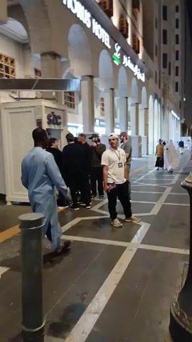 The video shows a group of people standing outside of a hotel at night. The hotel is called "Horus Hotel," and there is an STC booth nearby. The people are mostly dressed in casual clothing, and some are wearing traditional Muslim attire. They appear to be talking to each other. The ground is paved with a striped pattern. The lighting is dim, suggesting it is late in the evening. There are other pedestrians walking in the background.