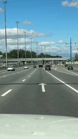 The video shows a forward-facing view from a vehicle driving on a multi-lane highway. The highway has a light gray asphalt surface with white lane markings. Tall, slender light poles with multiple lamps are visible along the right side of the highway, spaced at regular intervals. Trees and foliage line the left side of the highway, beyond a concrete barrier. In the distance, an overpass with multiple lanes of traffic can be seen. Several vehicles, including cars and a truck, are driving on the highway in the same direction as the camera. The sky is bright blue with scattered white clouds. The overall scene depicts a typical highway driving environment on a clear day.