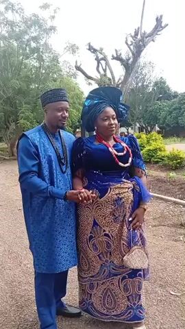 In this video, a couple is posing for a photo outdoors. The woman is wearing a vibrant blue traditional Nigerian outfit, complete with a matching headwrap and elaborate jewelry. The man is dressed in a blue traditional Nigerian attire, including a matching cap. They are standing close together, holding hands, and looking towards the camera with smiles.

The setting appears to be a park or a garden, with lush green trees and bushes visible in the background. The ground is covered with gravel and dirt. The lighting suggests it's daytime, possibly morning or afternoon, with soft, diffused light indicating an overcast or partly cloudy sky. The overall atmosphere is festive and celebratory, suggesting the couple might be attending a special occasion like a wedding or cultural event. There is no visible text in the video.