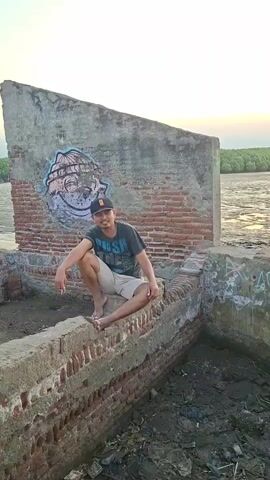 A man wearing a black baseball cap, a dark t-shirt with white lettering, and light-colored shorts sits on a brick wall. He is barefoot and holds a cigarette. Behind him is a graffiti-covered wall and a body of water. The scene appears to be outdoors, possibly a coastal area, with some vegetation visible in the background. The video pans to show a wider view of the area, revealing more people and several motorcycles parked along a pathway. Another man is seen sitting on a wall in the foreground, looking out at the water. The sky is overcast.