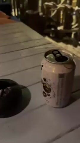 The video depicts two women seated at a light blue wooden table in an outdoor or semi-outdoor setting. Both women are looking at their phones. On the table, there is an open can of a pink drink, a dark ashtray with cigarette butts, a green lighter, a bottle of water, a small wooden tray with green food items, and a notebook. The women are sitting in front of a wooden fence-like structure, with greenery visible in the background. The overall lighting suggests it is evening or a dimly lit area.