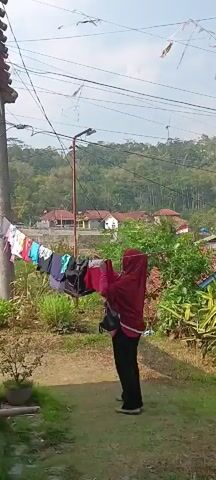 A person wearing a red hijab and black pants is hanging clothes on a clothesline outdoors. The clothesline is strung between a pole and what appears to be the side of a building. Behind the person and the clothesline, there are trees, hills, and houses in the distance. The sun is shining, casting shadows on the ground. The person is holding a red garment and appears to be in the process of hanging it. The ground is covered in dirt and grass. There are also some potted plants in the foreground. The overall scene is rural and domestic.
