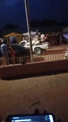 The video captures an outdoor scene at dusk or night. Several people are visible, some standing and interacting near a dark-colored car parked behind a fence. A white car is also parked in the background. Structures resembling small shops or stalls are present. The ground appears to be dirt or gravel, with some scattered debris. The sky is dark, indicating it is late evening or early morning. The lighting is dim, and some areas are in shadow. The camera is held at an angle, and the focus shifts occasionally. In the foreground, a phone screen is partially visible.