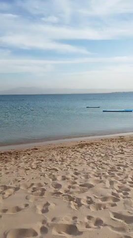 The video depicts a beach scene with a sandy shore in the foreground. The sand appears to be light brown and undisturbed, with various footprints scattered across the surface. The sea stretches out to the horizon, exhibiting a calm, light blue surface. Several floating platforms, appearing as dark rectangles and light blue rectangles, are visible in the water near the shoreline. The sky above is a mix of light blue and wispy white clouds. In the distance, a faint silhouette of land or hills can be seen along the horizon. The overall impression is that of a calm and serene beach setting.