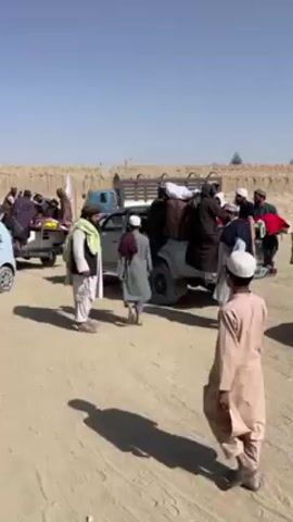 The video shows several people standing around and sitting in the back of a pickup truck in a dusty, dry, and sunny environment. The people, mostly men, are wearing traditional clothing. There are a few cars parked nearby. Some of the men are waving and appearing to be celebrating or preparing for a procession. A young boy in a tan robe walks away from the camera. There is a long wall behind the vehicles, presumably made of mud or clay.