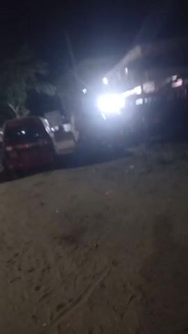 The video depicts a street scene at night in Mbribit, Nigeria. The foreground shows an unpaved area of dirt or sand alongside an asphalt road. Several vehicles are parked on the side, including a red car and a white van. In the background, artificial lights, likely from streetlights and a building or another vehicle, illuminate the dark surroundings. A vehicle with visible taillights briefly drives past on the road. No people or discernible text are visible in the video.