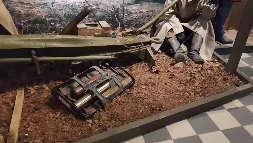 The video shows a diorama in a museum exhibit. In the foreground, there is a display case with what appear to be brass shells in a metal tray. Behind the tray, there is a pile of brown leaves and dirt, representing the ground. A mannequin dressed in a military uniform, including a trench coat and boots, is seated in the dirt, holding a telephone to its ear. The mannequin is positioned behind a green military artifact that resembles a weapon or equipment piece, with a long metal rod extending from it. The background of the diorama features a painted mural depicting a forest scene with other figures in military attire, suggesting a wartime setting. The floor in front of the diorama is tiled with a black and white checkered pattern.