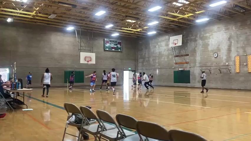 A basketball game is taking place in an indoor gymnasium. The court is made of polished wood with green and orange lines. There are several players on the court, all wearing athletic clothing. Some players are actively engaged in the game, dribbling or passing the ball, while others are positioned on the court. Spectators are seated in chairs to the left of the frame, with a table beside them holding drinks and trophies. The gymnasium has a high ceiling with exposed beams and fluorescent lighting. Two basketball hoops are visible on the far wall. A scoreboard is mounted above one of the hoops, displaying game information. The walls are made of grey concrete blocks.