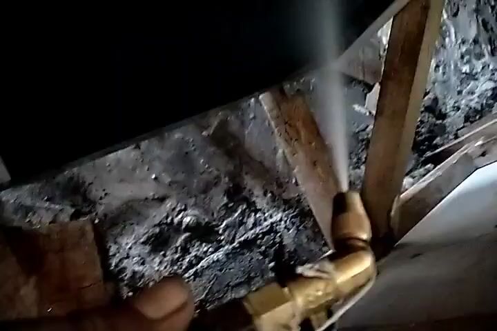 A close-up, shaky video shows a person's hand holding a brass plumbing fitting that appears to be connected to a hose or pipe. Water is spraying forcefully from the fitting, creating a mist or spray. The background is dark and appears to be an unfinished or cluttered space, with wood and possibly concrete visible. The focus is on the spraying water and the fitting.