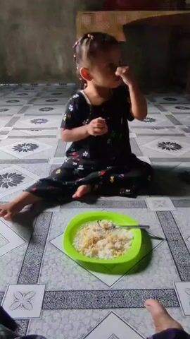 A young child with dark hair pulled into two pigtails, wearing a black outfit with a floral print, sits on a tiled floor with a green plastic plate of rice and what appears to be shredded cheese or some other topping in front of them. The child is holding their hand to their mouth and looking towards the camera. The floor has a gray and white patterned tile design. In the background, a woven mat is visible on a table, and a wall with a concrete finish is present. The lighting suggests an indoor setting.