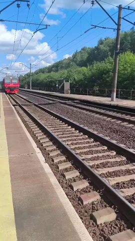 A red and black train approaches on a set of train tracks. The train is on the left side of the image. The train tracks are in the foreground and middle ground. There is a platform to the left of the train tracks. The train has lights on its front. There are power lines above the train tracks and to the left of the image. Trees and vegetation are visible behind the train tracks and to the right of the image. The sky is blue with some clouds. The image appears to be taken from a stationary position on the platform.