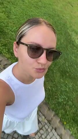 The video begins with a close-up selfie of a woman with blonde hair and sunglasses, wearing a white tank top and white pants. She is smiling and walking on a cobblestone path. The camera pans to reveal a young girl in a white t-shirt and blue shorts, holding a pink toy rake, standing on the grass next to the path. The background shows a green lawn, a tree, and a building. The woman continues to walk, and the camera shows her walking beside the girl, who is walking in the same direction. The scene appears to be in a park or a residential area with well-maintained landscaping.