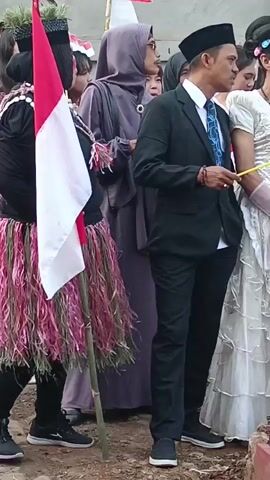 A group of people are gathered outdoors. In the foreground, a person in a black suit and cap holds a flag that is red and white, with a bamboo pole. To their right, a person in a white, ruffled dress, long white gloves, and sunglasses holds a small red and white flag. Behind them, a person in a purple hijab and glasses stands next to another person in a black suit. To the right of the person in the white dress, a person in white robes and a bald head stands, and further to the right, a child in a costume with red and white elements and a green balloon is visible. The ground appears to be dirt and rocks, with some red bricks or tiles visible in the lower right corner. The people are interacting, with some looking at each other and some looking away. The overall atmosphere seems to be a gathering or event.