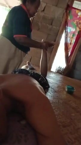 An elderly woman massages the raised leg of a person lying on a mat. The woman stands to the left of the person, her back to the camera. She wears a dark blue collared shirt with a red band around the sleeve cuffs and a tan skirt. The person is lying face-down on a woven mat with their head to the left. They are wearing dark shorts. There is a small container with a green substance to the right. The room has exposed brick walls and a partially visible window with a patterned fabric.