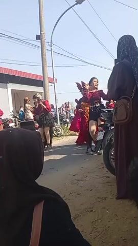 The video captures an outdoor scene with a group of people, seemingly in a street or public area. The primary focus is on a young woman in the center, dressed in a red top with voluminous sleeves, black shorts, and black boots. She is performing a dance routine with energetic movements. Behind her, other individuals are also present, some in similar costumes, including another woman in a red outfit with a flowing skirt. To the right, a woman wearing a brown hijab and dress is observing the performance. In the foreground, parts of people are visible, also watching. The environment appears to be a somewhat dusty, unpaved area with buildings and parked motorcycles in the background. A utility pole with wires runs through the frame. The lighting suggests it is daytime.