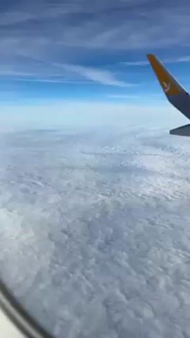 The scene is a view from an airplane window. The sky is a bright blue with sparse streaks of wispy clouds. Below, a dense layer of white clouds stretches out to the horizon. The wing of the plane, bearing a yellow logo, is visible in the upper right corner of the frame. The plane is in motion, presumably in flight.