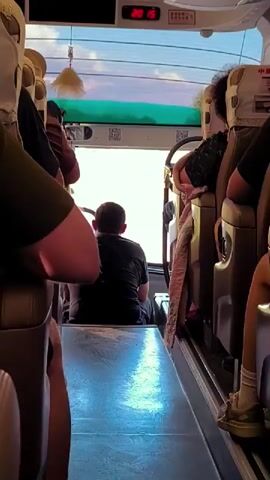 The video begins inside a bus, showing the aisle from a low angle. Passengers are seated on either side. In the center of the frame, a person in a dark t-shirt is seated facing away from the camera. The bus has white headrest covers with some text and images on them. Sunlight streams in from the front of the bus, creating a bright, overexposed view. As the video progresses, the perspective shifts to show the view out of the bus windows. The bus appears to be moving, as the scenery outside is blurred. The exterior view shows a clear sky with some clouds, trees, and buildings in the distance. An exit sign is visible on the upper right side of the window. The video ends showing the blurred scenery outside the bus.