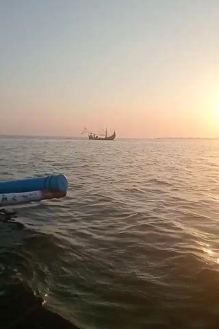 The video shows a body of water with small waves. The sky is clear and a bright sun is visible on the right side of the frame, setting or rising. In the distance, a small boat with a few people on board is visible. The boat appears to be moving. In the foreground on the left, a blue and white cylindrical object is partially visible, likely part of the vessel from which the video is being recorded. The overall scene suggests a calm day on the water.