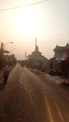 The video shows a street scene with a mosque on the right side of the road. The sun is setting, casting a warm glow over the scene. Several people are riding motorcycles on the road, moving away from the camera. One person is riding a motorcycle towards the camera. There are flags on the side of the road, possibly indicating a celebration or national holiday. The road appears to be paved and in good condition. The buildings on the right side of the road are simple, with one building appearing to be a shop or stall. The overall atmosphere of the video is peaceful and serene.