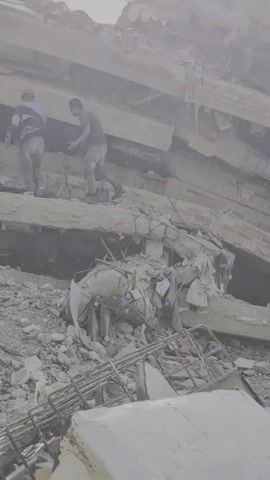Two people are visible in the foreground, one wearing a hard hat. They are standing on a pile of rubble, which appears to be the remains of a collapsed building. Concrete slabs and rebar are scattered everywhere. In the background, more people, some in uniform, are seen navigating the debris. The scene suggests a disaster or construction site where a building has collapsed, and rescue or recovery efforts may be underway. The lighting is hazy, possibly due to dust or atmospheric conditions.