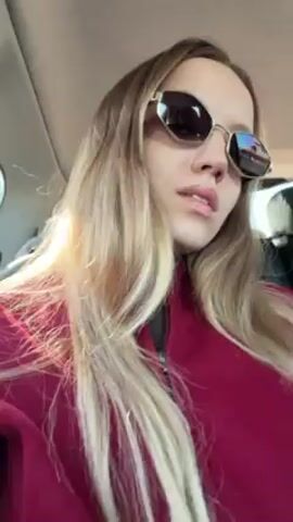 A young woman with long blonde hair is in a car, wearing a red jacket and gold-framed sunglasses with dark lenses. She looks at the camera and adjusts her sunglasses with her left hand. The car's interior is visible in the background.