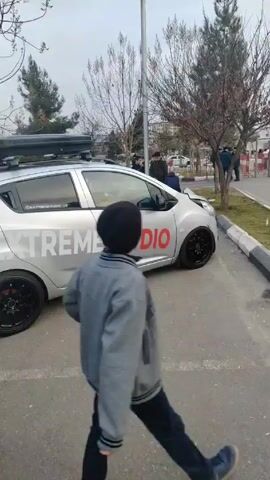 The video shows a light silver car with the words "EXTREME AUDIO" in large red letters on its side. The car has black wheels and a roof rack, and a large custom-built speaker system attached to the back, with a box containing additional speakers on the ground nearby. Several people are standing around the car, including a young boy who is taking pictures with his phone. One man in a gray jacket is looking toward the camera, and others are milling about. The scene is an outdoor parking area, with bare trees and buildings in the background. The video appears to be documenting the sound system installed on the car.