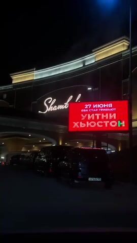 The video shows a nighttime view of a building with a large illuminated sign that reads "Shambala." In front of the building, there is a queue of cars, with some of them driving under an archway. A red digital billboard is visible on the right side of the screen, displaying text in Russian. The lighting suggests an entertainment venue, possibly a casino or theater. The overall atmosphere is one of an active evening with traffic and illuminated signage.