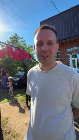 A person is filming an outdoor scene on a sunny day. In the foreground, a person in a white t-shirt and dark hair is speaking to the camera. The camera then pans right to show a fence and a metal archway adorned with red heart-shaped balloons. Beyond the fence, a black car is parked, and several people are visible in the background, including a woman in a black top and blue jeans, another person near a white car, and a small child in a blue shirt and shorts. The sun is brightly visible in the top left of the frame. The ground is dirt and grass.