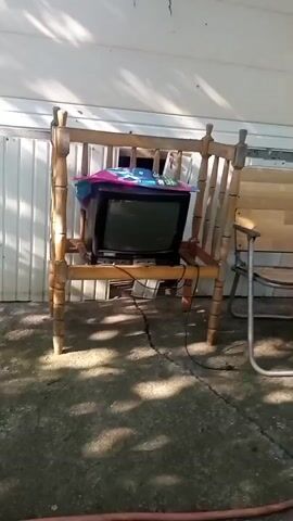 A vintage television is perched on a wooden chair outside next to a white-paneled building. The television is covered with colorful fabric on top. A power cord extends from the television to the ground. To the right of the television chair is a folding metal chair with a wooden seat and back. Further to the right, a small green stool is visible. The ground is a concrete patio with visible cracks. Sunlight casts shadows on the ground, suggesting it is daytime. The scene appears to be in an outdoor, possibly backyard, setting with trees and foliage visible in the background. The overall impression is one of a weathered, somewhat neglected outdoor area.