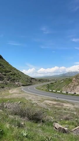 The video shows a scenic landscape in Armenia. A winding road cuts through a valley between two hills, surrounded by grassy terrain. The sky is a clear blue with some scattered clouds. A van and a car are parked on the side of the road. A man in a gray shirt walks towards the vehicles. Power lines run along the road.
