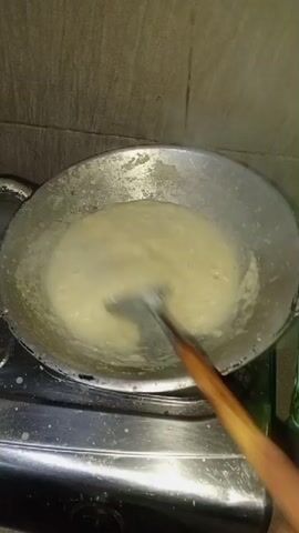 A person's hand is visible on the right side of the frame, holding a wooden-handled utensil. The utensil is being used to stir a pale yellow, semi-liquid substance in a round, metallic pan. The pan is placed on a gas stove, with its burner visible beneath the pan. Steam is rising from the substance in the pan. The background consists of a tiled wall.