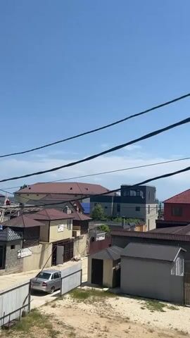 A video pans right across a residential area with a clear blue sky and a few clouds. Power lines stretch across the top of the frame. In the foreground, there is a dry, sandy patch with some weeds. Behind this, there are several low-lying buildings with metal and tiled roofs, some with fences. A silver car is parked in a driveway on the left. Further back, there are larger houses with red and brown roofs, and a modern grey building with large windows. In the distance, the blue sea is visible, with a faint line of trees or vegetation along the shore. The overall impression is of a sunny day in a coastal town or village.